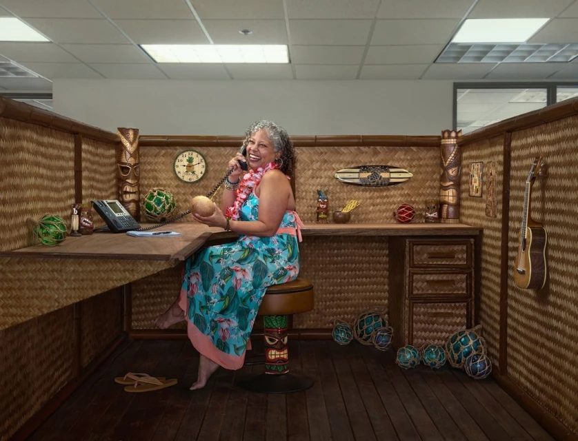 Smiling woman in floral dress sits at a desk in an office cubicle decorated with tiki statues and Hawaiian-themed items, talking on the phone.